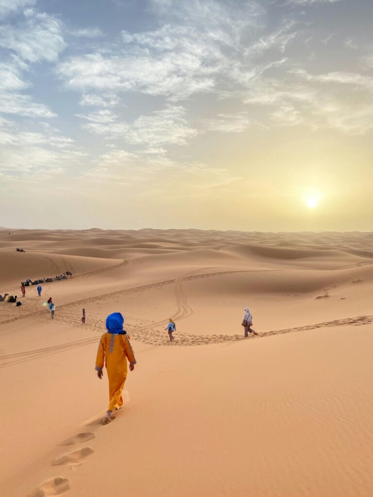 Travelers walking through the vast dunes of Merzouga Desert during a stunning sunset in Morocco.